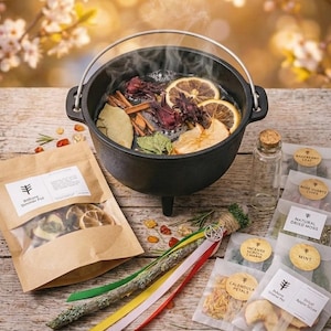 May include: A black cast iron simmer pot with a silver handle, filled with dried fruit and herbs. A kraft paper bag labeled "Bottane Simmer Pot" and small packets of herbs and botanicals are arranged on a wooden surface.