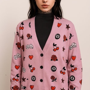 May include: A light pink cardigan sweater with a V-neck and black buttons. The sweater is patterned with red cherries, roses, hearts, stars, dice, and eight ball graphics. The cardigan is a button-up style.