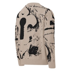 May include: Beige sweater featuring a black keyhole and abstract brushstroke design. The long-sleeved sweater has a crew neck and ribbed cuffs and hem. The back of the sweater is visible, showcasing the bold, artistic pattern.