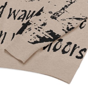 May include: Close-up of a beige sweater with black text and abstract designs. The sweater has a ribbed cuff and hem. The text appears to be a partial phrase, with the word "way" visible.