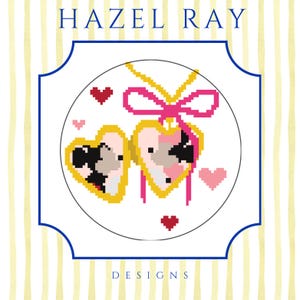 May include: A cross-stitch design featuring two heart-shaped lockets with pink and black details, accented by a pink bow and small hearts. The design is set against a white background with the text "HAZEL RAY DESIGNS".