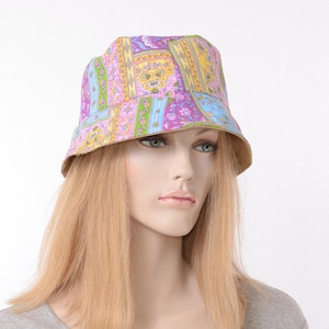 May include: A colorful bucket hat with a paisley pattern in shades of purple, yellow, and blue. The hat has a tan-colored brim and is worn by a mannequin. The hat's design features floral and geometric elements.