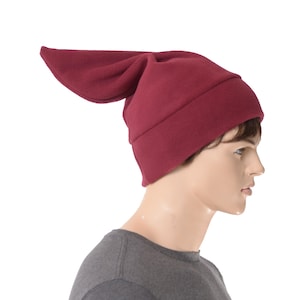 May include: A burgundy-colored hat with a long, pointed flap. The hat is made of a soft material and has a rounded crown. The hat is worn by a mannequin. The hat is a unique and stylish accessory.
