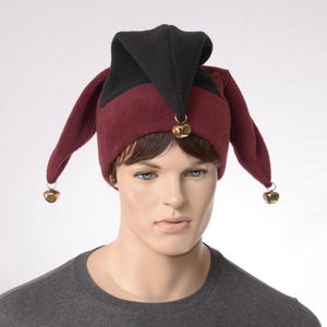 May include: A jester hat featuring a black pointed top section and a deep burgundy base with two long, drooping side flaps. Each of the three points of the hat is adorned with a small, metallic gold-colored bell attached by a circular ring.