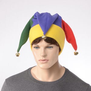 May include: A multi-colored jester hat featuring four distinct fabric points in green, purple, yellow, and red. The hat is constructed from soft fleece material and includes small, metallic gold-toned bells attached to the tips of the points.