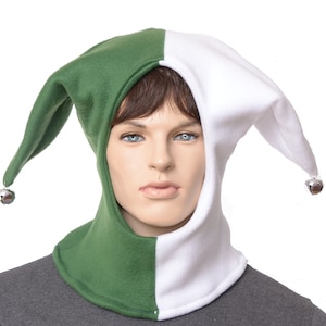 May include: A two-toned jester hat featuring a split design with one side in forest green and the other in white. The hat has two long, pointed ear-like flaps that extend downwards, each finished with a small, metallic silver jingle bell.
