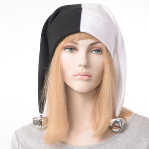 May include: A two-toned jester hat featuring a split design with one side in solid black and the other in solid white. The hat has a long, pointed shape that hangs down on both sides, each finished with a large, shiny silver-toned metal jingle bell.
