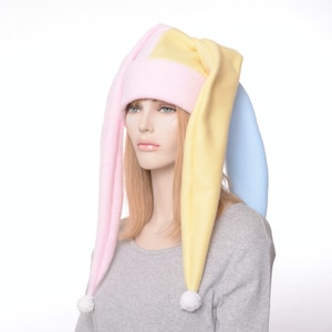 May include: A pink, yellow, and blue fleece hat with long, pointed ears. The hat has white pom-poms on the ends of the ears.