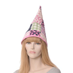May include: A patchwork fabric hat with a pointed top. The hat has a pink brim and is made of various floral and checkered patterned fabrics in pink, green, and navy blue. The hat is worn by a mannequin with blonde hair.