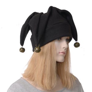 May include: A black jester-style hat featuring three distinct points that hang down. Each of the three points is finished with a small, round, metallic brass-coloured bell attached by a ring. The hat is constructed from a solid black fabric material.