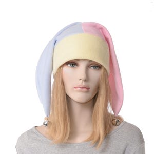May include: A multi-colored jester hat featuring a pale yellow headband, a light blue side panel, and a light pink side panel. The hat has two long, drooping ear-like flaps, each tipped with a small metallic jingle bell at the bottom edge.