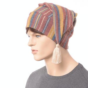 May include: A striped, colorful hat with a tassel. The hat features vertical stripes in shades of red, yellow, and blue. A cream-colored tassel hangs from the side. The hat is worn by a mannequin.