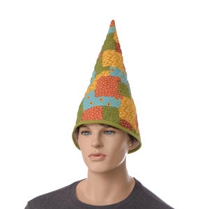 May include: A tall, cone-shaped hat featuring a quilted patchwork design with various fabric squares in shades of green, orange, yellow, and light blue. The hat has a solid green fabric trim along the bottom edge and a pointed top structure.