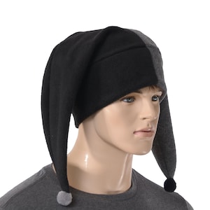 May include: A two-toned jester-style hat featuring a split design with one side in solid black and the other in dark charcoal gray. The hat has two long, pointed earflaps that hang down, each finished with a small, round pom-pom at the tip.
