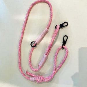 Pode incluir: Alça de corda rosa e branca com glitter e fechos de metal preto. A alça tem uma seção com nós e uma seção enrolada em um rosa mais claro. Projetado para um telefone ou outro item pequeno.
