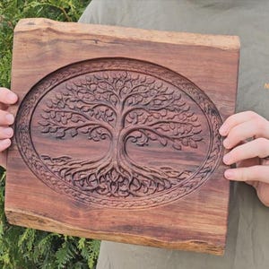 May include: A square wooden carving with a tree of life design. The tree is carved within an oval frame, with detailed roots and branches. The wood is a rich, dark brown colour and has a natural edge. The carving is approximately 30 cm square.