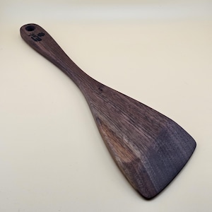 May include: A dark brown wooden spatula with a long handle and a wide, flat head. The handle features a circular hole at the top for hanging and a small, dark stamped logo that reads Crafted by Hand. The wood grain is visible throughout the surface.