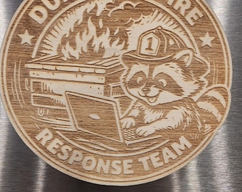 May include: Circular wooden emblem with the text "DUMPSTER FIRE RESPONSE TEAM" around the edge. The center features a raccoon wearing a firefighter helmet, working on a laptop, with a dumpster on fire in the background.