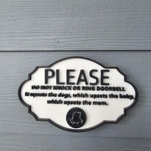 Puede incluir: Un letrero blanco y negro con el texto "PLEASE DO NOT KNOCK OR RING DOORBELL It upsets the dogs, which upsets the baby, which upsets the mom." El letrero tiene un icono de campana y está montado sobre una superficie de madera gris.