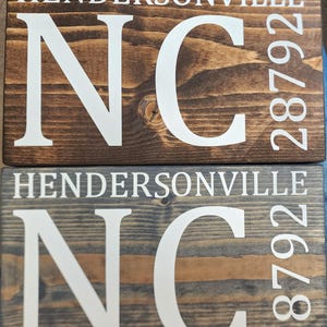 Może przedstawiać: Dwa drewniane znaki z napisem "HENDERSONVILLE NC 28792" w kolorze białym. Jeden znak jest bejcowany na brązowo, drugi na szaro. Znaki są wykonane z drewna i mają rustykalny wygląd.