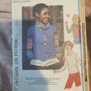 Peut inclure: Patron Simplicity 8126 pour petites filles et rondes. L'enveloppe du patron présente une photo d'une jeune personne portant un gilet bleu et tenant un livre. Le texte sur l'enveloppe comprend "One Pattern Un Patron" et "Facile-A-Coudre/Simple-To-Sew".