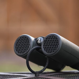 May include: A pair of black binoculars with a textured, honeycomb-like design on the lenses. The binoculars are sitting on a metal grate.