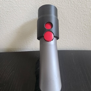 Puede incluir: Un accesorio de aspiradora negro y gris con un botón rojo. El accesorio tiene un cepillo en la parte inferior.