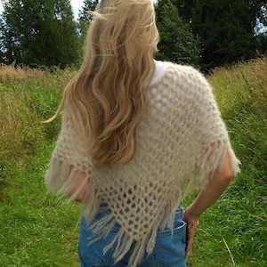 Pode incluir: Um xale bege de tecido aberto com franjas. O xale é usado sobre uma blusa branca e jeans azuis. A imagem é tirada ao ar livre com um fundo de grama verde e árvores.