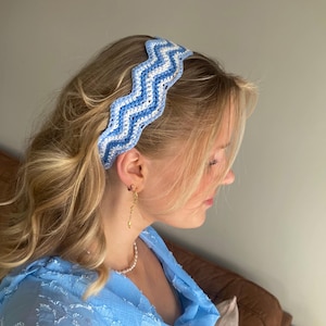 Puede incluir: Una diadema de ganchillo azul y blanca con un patrón ondulado. La diadema se lleva en la cabeza. La imagen también muestra una blusa azul, un collar de perlas y pendientes dorados.