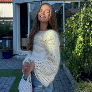Puede incluir: Un bolero de crochet color crema con mangas acampanadas se usa sobre una camiseta blanca. El bolero tiene un diseño abierto y aireado. La persona lleva vaqueros azules y sostiene un pequeño bolso de crochet a juego. El fondo incluye una casa y vegetación.