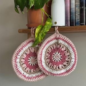 Puede incluir: Dos agarraderas redondas tejidas a mano con un patrón circular estilo mandala en tonos rosa empolvado, crema y marrón claro. Cada agarradera tiene un pequeño lazo trenzado en la parte superior para colgar y una textura de hilo grueso.