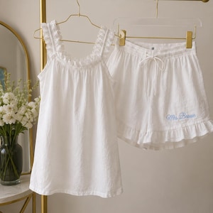 May include: A white two-piece pyjama set consisting of a sleeveless top with ruffled straps and matching shorts with a ruffled hem. The shorts feature the embroidered text Mrs. Brown in light blue script on the lower right side.