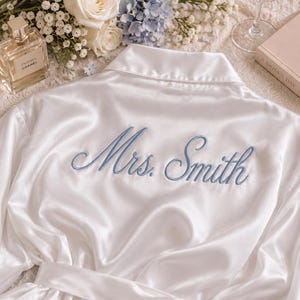 May include: A white satin robe featuring long sleeves, a collar, and a tie waist. The back of the robe is embroidered with the text Mrs. Smith in a dusty blue script font, centered across the upper back area of the garment.