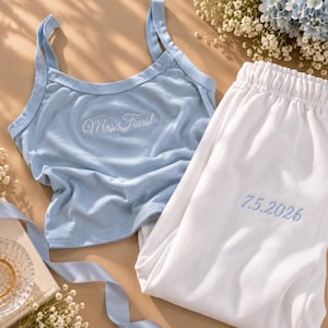 May include: A light blue tank top with white embroidery reading Mrs. Faust and white pajama pants with light blue embroidery reading 7.5.2026. The set features a scoop neckline, thick straps, and an elastic waistband on the pants.