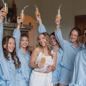 May include: A group of individuals wearing matching light blue and white vertical striped button-down pajama shirts and shorts, with one individual in a white tank top, all holding tall champagne flutes filled with orange juice and pink straws.
