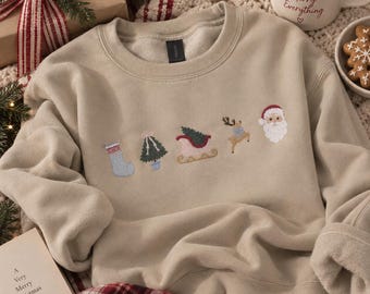 Könnte beinhalten: Ein beigefarbenes Sweatshirt mit Rundhalsausschnitt und einer Reihe kleiner gestickter Weihnachtsmotive, darunter ein hellblauer Strumpf, ein grüner Baum, ein roter Schlitten, ein braunes Rentier und ein Weihnachtsmannkopf auf der Brust.