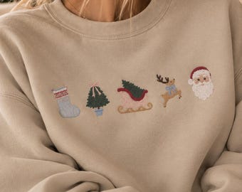 Könnte beinhalten: Ein beigefarbenes Sweatshirt mit Rundhalsausschnitt und einer horizontalen Reihe aus fünf gestickten Weihnachtsmotiven: ein hellblauer Strumpf, ein grüner Baum im Topf mit rosa Schleife, ein rot-grüner Schlitten, ein braunes Rentier und der Weihnachtsmann.