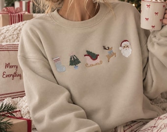 Può includere: Una felpa beige a girocollo con una fila orizzontale di icone natalizie ricamate, tra cui una calza blu, un abete verde, una slitta rossa, una renna marrone con un fiocco blu e il volto di Babbo Natale con un cappello rosso.