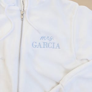May include: White zip-up hoodie with the text "mrs. GARCIA" embroidered in light blue. The hoodie features a drawstring hood, a full-length zipper, and long sleeves. The brand label "BELLA+CANVAS" is visible on the inside of the collar.