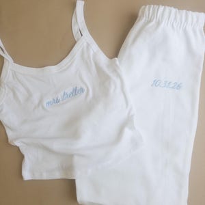 May include: White pajama set with a camisole top and trousers. The top has thin straps and the embroidered text "mrs. treller" in light blue. The trousers have an elasticated waistband and the embroidered date "10.31.26" in light blue.