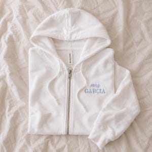 May include: A white zip-up hoodie featuring a hood with matching drawstrings and a silver-toned metal zip. The left chest area displays the embroidered text "mrs. GARCIA" in a light blue script and block font combination.