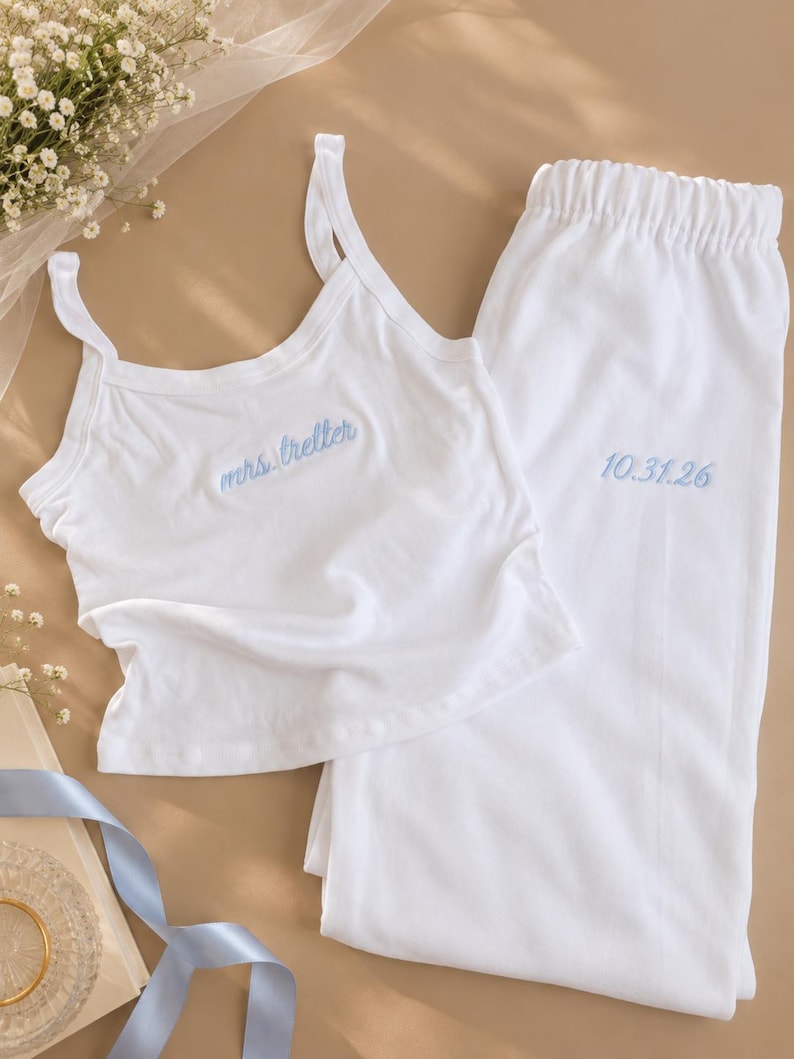 May include: A white pajama set consisting of a tank top and pants. The tank top features the embroidered blue text "mrs. breller" on the chest. The pants have an elastic waistband and the embroidered blue date "10.31.26" on the upper leg.