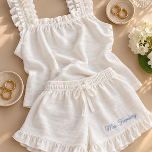 May include: A white two-piece lounge set consisting of a sleeveless top with ruffled shoulder straps and matching shorts with a ruffled hem. The shorts feature a drawstring waist and the embroidered blue text Mrs. Framberg.
