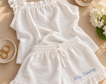 Puede incluir: Un conjunto de descanso blanco de dos piezas que consta de una parte superior sin mangas con tirantes con volantes y pantalones cortos a juego con dobladillo con volantes. Los pantalones tienen cordón y el texto Mrs. Framberg bordado en azul.