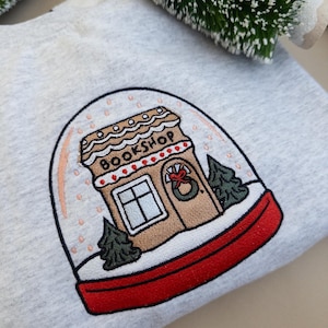 May include: A light gray sweatshirt featuring an embroidered snow globe design. The snow globe contains a gingerbread house with the word "BOOKSHOP" above the door. The base of the snow globe is red, and the design includes small green trees.