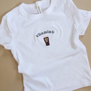 May include: White t-shirt with the embroidered text "Vitamin G" above a graphic of a dark stout in a glass. The shirt is made of a soft material and has short sleeves.