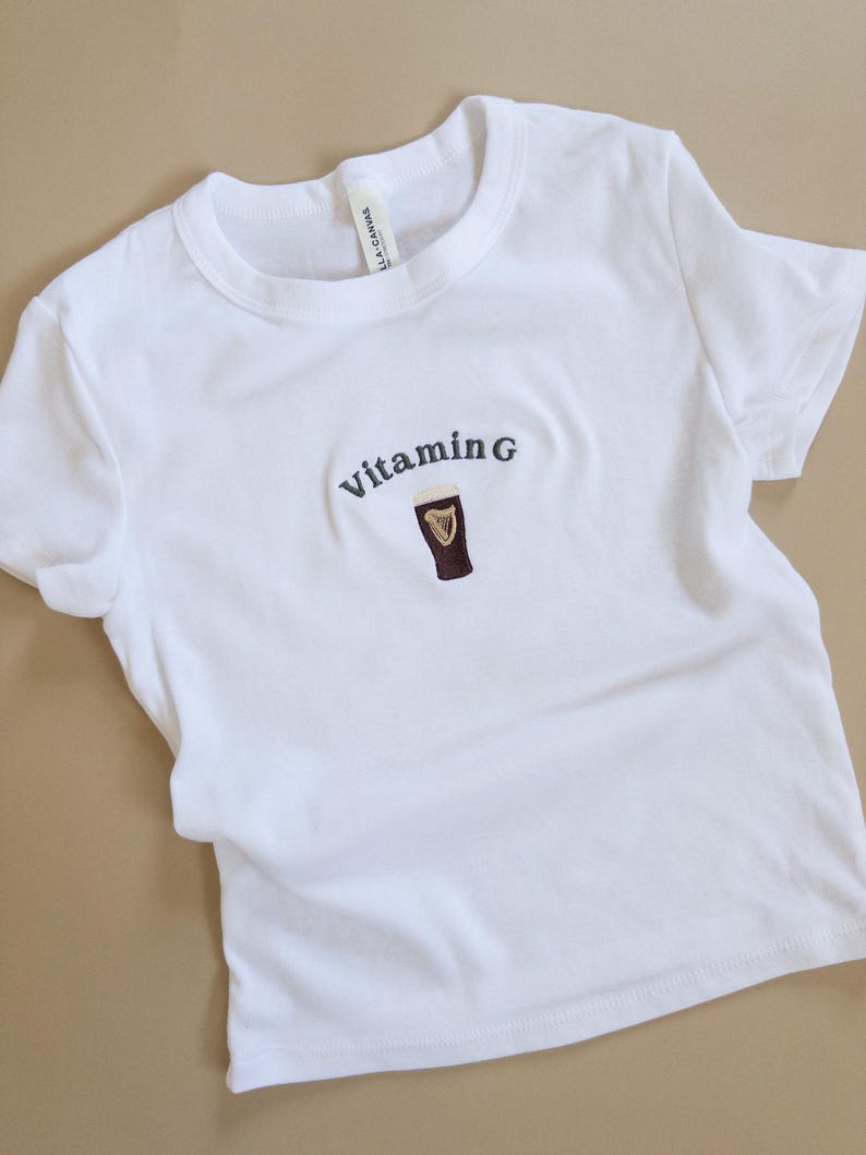 May include: White t-shirt with the embroidered text "Vitamin G" above a graphic of a dark stout in a glass. The shirt is made of a soft material and has short sleeves.