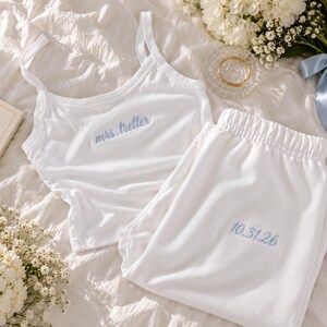 May include: A white pajama set consisting of a tank top and elastic-waist pants. The tank top features the embroidered blue text "mrs. tretter" on the chest, and the pants feature the embroidered blue date "10.31.26" on the upper leg area.