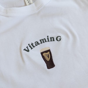 May include: White t-shirt with the embroidered text "Vitamin G" above a brown pint of beer with a gold harp logo. The shirt is made of a soft material and has a crew neck.