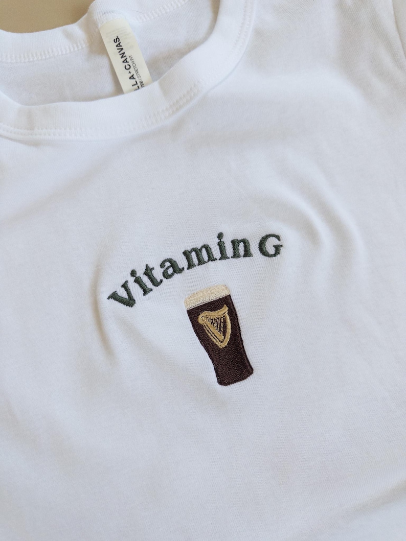May include: White t-shirt with the embroidered text "Vitamin G" above a brown pint of beer with a gold harp logo. The shirt is made of a soft material and has a crew neck.